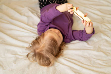 Sweet little girl gen Z using online app on smartphone, browsing internet, playing videogames, taking selfie, making call, watching cartoons, resting on bed in bedroom at home.の写真素材