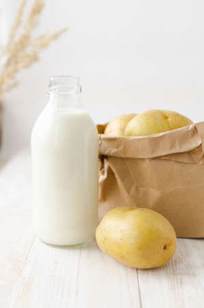 Potato milk in a glass bottle on a white wooden background. New, young potatoes in a kraft paper bag on the tableの写真素材