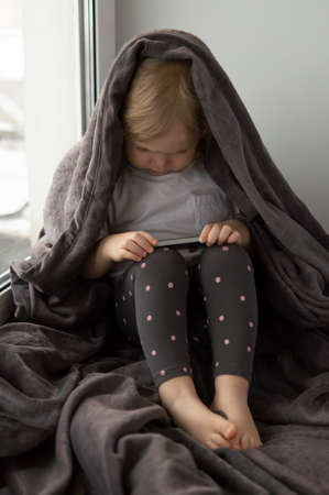 Vertical image of a little caucasian girl sitting on window, wrapped in a soft blanket and using her mobile phone.の写真素材