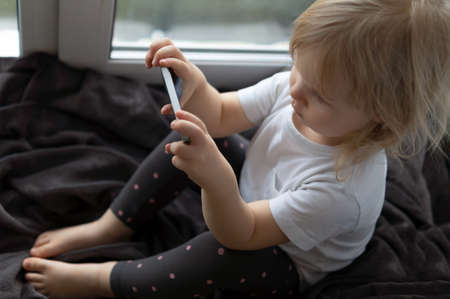 Top view of a little girl sitting on the window in a gray blanket and watching cartoons on her smartphone.の写真素材