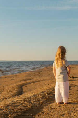 Little cute girl with blond hair standing on the seashore barefoot at sunset and looking into the distance. Local travel trend, authentic tourism.の写真素材