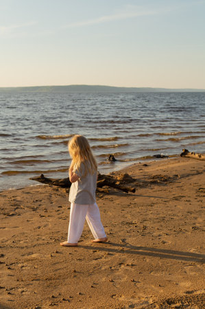 Cute little girl with blonde hair walking barefoot on the beach at sunset on a summer evening. Local travel concept, authentic tourism.の写真素材