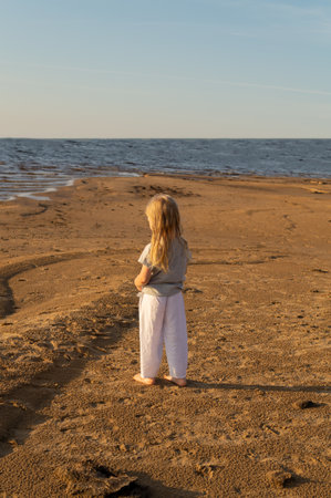 Adorable little girl is standing barefoot on the seashore at sunset on a summer evening. Carefree childhood, traveling with family.の写真素材
