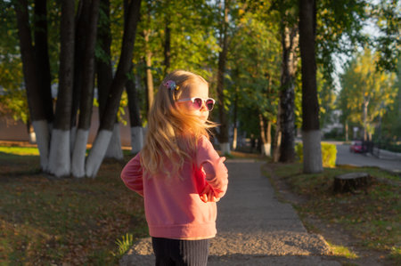 Charming little girl in glasses with long blonde hair is walking and having fun in the park in the autumn season at sunset, her hair is in motion.の写真素材
