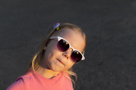 Funny little girl with blond hair in sunglasses standing on the street on a background of gray asphalt in the sunlight.の写真素材