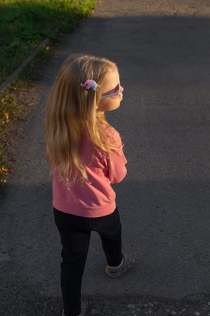 Beautiful little girl in sunglasses with long blonde hair walking in an autumn park at sunset.の写真素材