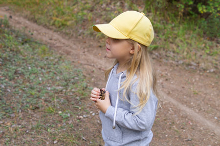 Little cute girl in a yellow cap with long blonde natural hair and casual clothes collecting cones on a walk in the autumn forest.の写真素材