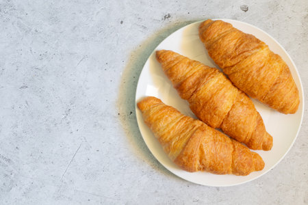 Three fresh croissants on a white plate on a light concrete background. Top view, flat lay, with copy space for your text on the leftの写真素材