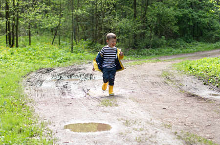 A happy little boy in a yellow jacket and rubber boots runs through the puddles and laughs. Happy childhood conceptの写真素材