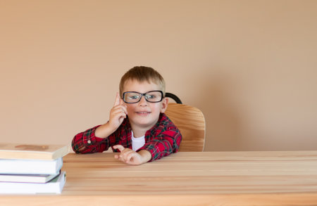 A little cheerful boy with glasses sits at a desk and points his finger at something. Back to school concept. Copyspaceの写真素材