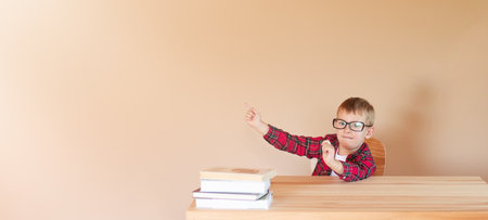 A little cheerful boy with glasses sits at a desk and points his finger at something. Back to school concept. Copyspaceの写真素材