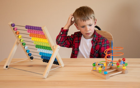 A little boy sits at the table and plays with wooden educational toys, scratches his head and thinks...