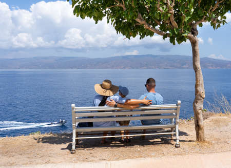Happy family sits on a bench in the shade of a tree and looks at the sea. Holidays and vacation at sea conceptの写真素材