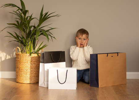 Little boy sits on the floor next to shopping bags. He is surprised and hugged his face in his hands. Black Friday and Cyber Monday concept. Mockupの写真素材