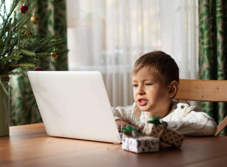 Little boy in a white sweater is sitting at a laptop and speaking with his grandparents by video call. Christmas preparationの写真素材