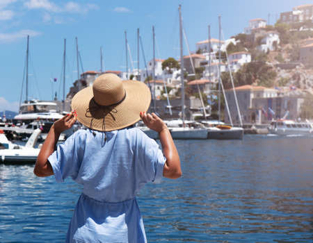 A young woman in a blue dress and a large straw hat stands with her back and looks at the sea bay