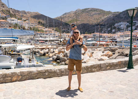 Father and son on the background of the port in the Greek city. The boy sits on dad's shoulders. International Father's Day.の写真素材