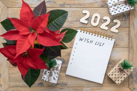 Notepad on the wooden background with the inscription - Wish list. Poinsettia in the pot, gift boxes and wooden numbers 2022の写真素材