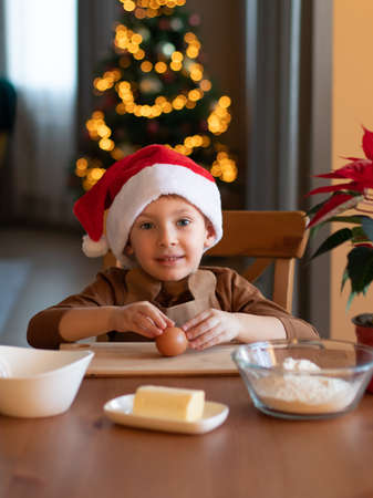 A little boy in an apron and Santa Claus hat prepares Christmas cookies. Christmas tree in the background.の写真素材