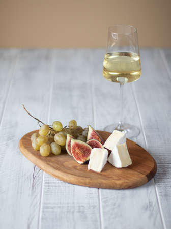 A glass of white wine and wine snacks - cheese, figs, grapes on a wooden board on a light background. Wine tasting, home gatherings and restaurant meetings.の写真素材
