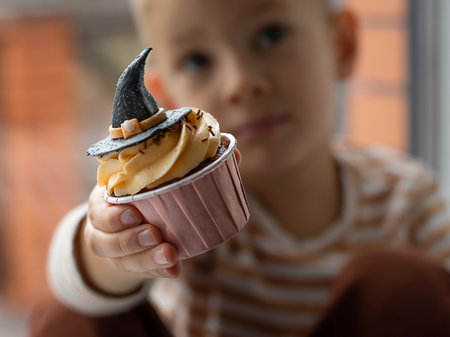 A little boy sits on the windowsill and eats a cupcake in the form of a witch's hat. Halloween concept.の写真素材