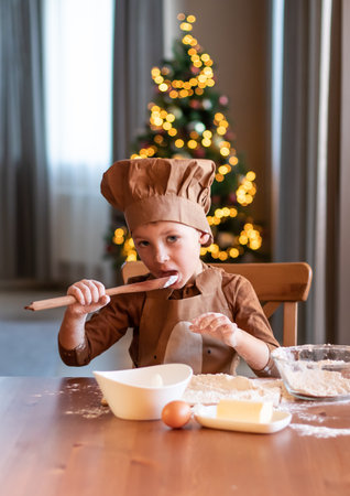 A little boy in an apron and a chefs hat prepares Christmas cookies. Christmas tree in the backgroundの写真素材