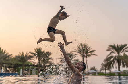 Dad and son are playing in the swimming pool. The father tosses the son up.