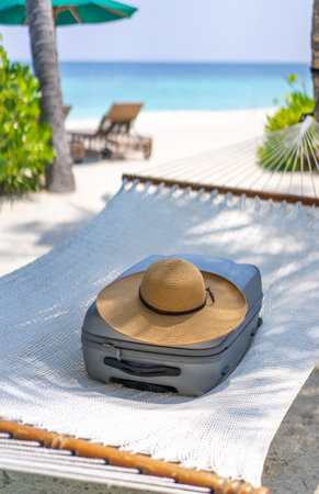 A suitcase and a straw hat lying on a hammock in the backdrop of the landscape of the Maldives....