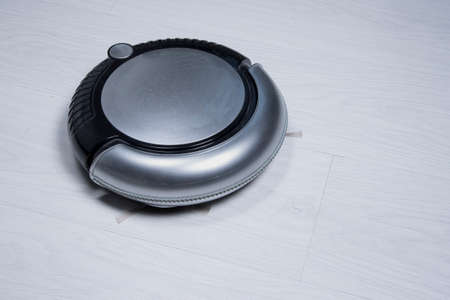 Black and silver robotic vacuum cleaner on white laminate wood floor. Smart home.