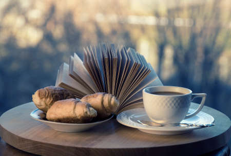 White coffee cup, croissants and brown open book on wooden table in cafe. Breakfast in coffee palace. Window view. Blurred outside background. Selective focus.の写真素材