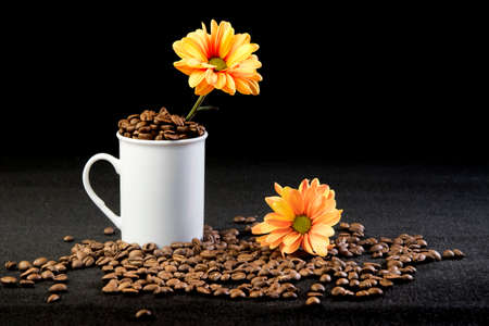 Coffee beans in white cup and bright orange flowers on black and gray background. Selective focus.