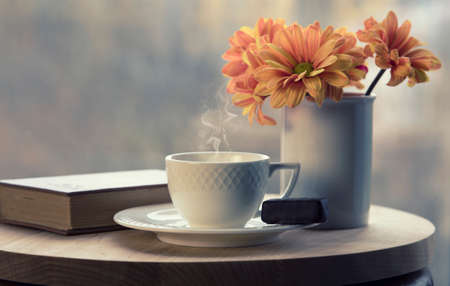 White coffee cup with smoke, vase with orange flowers and brown book on table in cafe. Late autumn and coziness concept. Selective focus.
