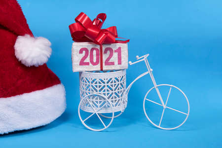 2021 text on gift box with red ribborn on bicycle and santa claus hat. Blue background. Christmas...