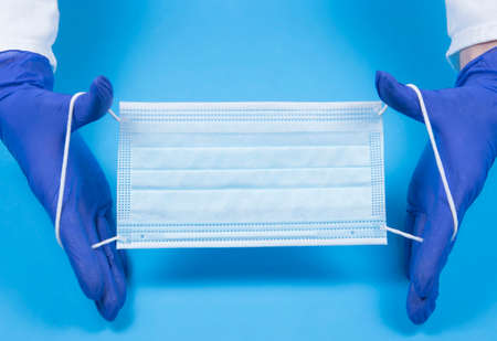 Doctor's hands holding medical mask on blue background top view with copy space. Hands in gloves...