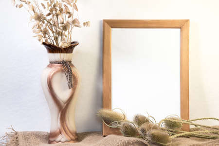 Portrait empty wooden frame mockup with dry grass and ceramic vase on sackcloth. White background....