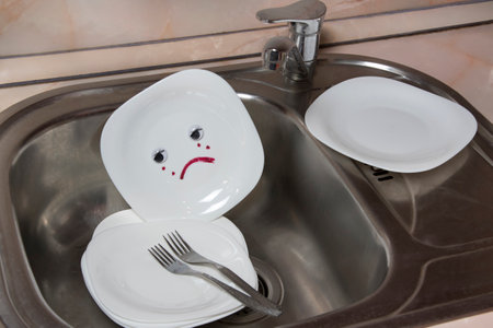 Poorly washed dishes in kitchen sink. White plate with sad emotion. Kitchen faucet and forksの写真素材