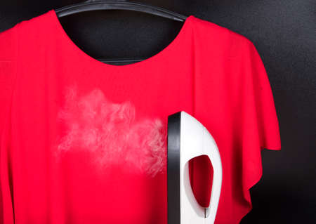 Portable home and garment steamer for clothes and red blouse on hanger on black background. Housework with stream ironの写真素材