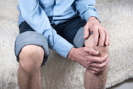 Cropped shot of elderly man sitting on sofa and suffering from pain in knee. Joint disease, arthritis or arthrosisの写真素材