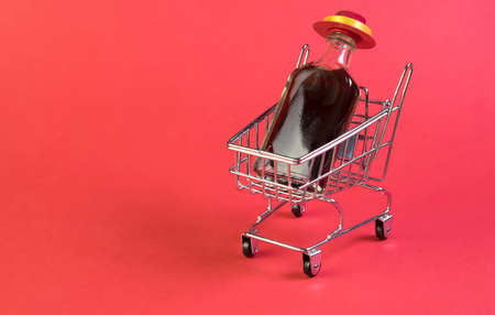 Small wine bottle with red hat in mini shopping cart on red background. Alcohol delivering concept