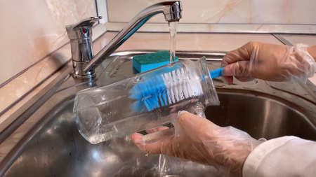 Woman washes glass jug in kitchen sink. Close up of pitcher, faucet and female hand in gloves. Water streaming. Brown backgroundの写真素材