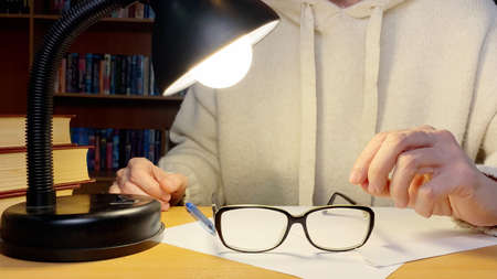 Middle-aged hands with glasses in library near lamp. Bookshelves on background.の写真素材