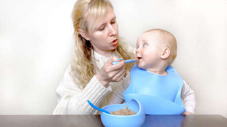 Mother feeding little kid with blue spoon. Baby eating cereal from bowl. Infant food, healthy eating concept. Child makes funny facesの写真素材