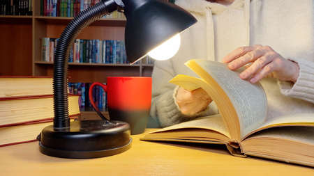 Middle-aged hands turning pages. Woman reading book in library near lamp. Coffee cap.の写真素材