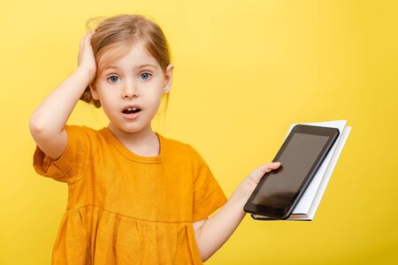A little blonde girl in a yellow dress on a yellow background with a book and laptop.