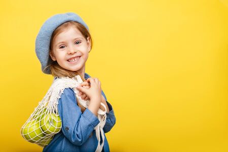 A little blonde girl with long hair in a blue dress and beret on a yellow isolated background. Child with apples.の写真素材