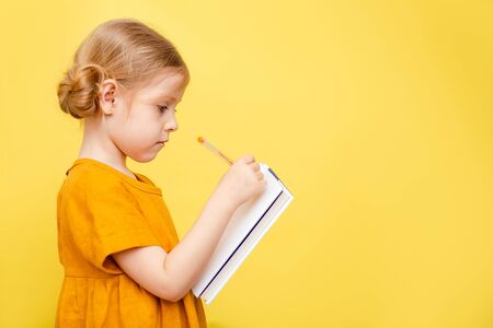 A little girl with light hair on a yellow background. Isolated. A child in a yellow dress makes notes on a book.の写真素材