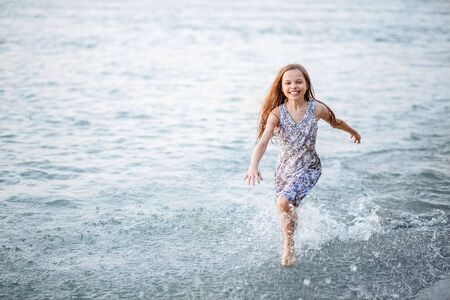 A happy young girl runs along the shore on the water and waves her hat. The girl is happy with the onset of summerの写真素材