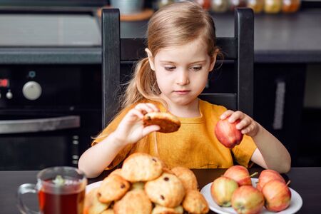 Healthy and harmful food during quarantine, self-isolation. The little blonde girl chooses to take an unhealthy cookie or Apple.の写真素材