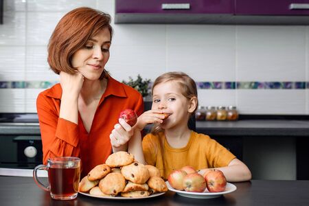 Difficult choice between healthy and unhealthy food during quarantine, isolation at home. Mother and daughter in the kitchen. A little girl eats delicious homemade cookies.の写真素材