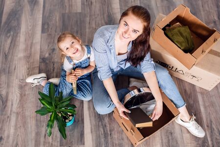 Mother and daughter sort things out after moving to a new house or apartment. A woman and a little girl sit on the floor among cardboard boxes and read a book.の写真素材